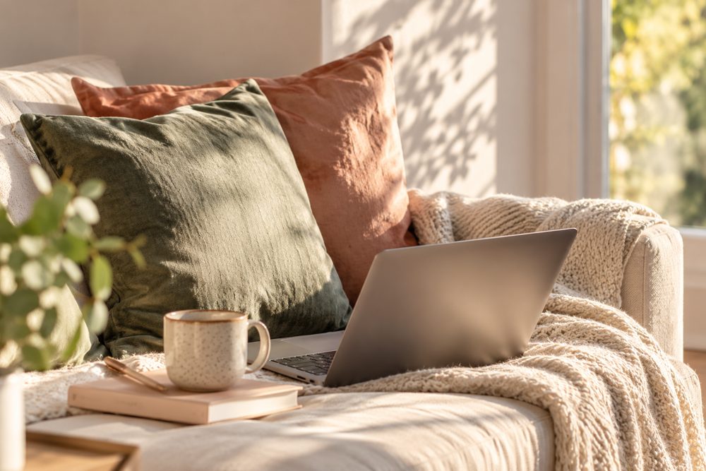 Sunlit sofa with warm terracotta and olive cushions, a cosy blanket and a laptop ready for an online session